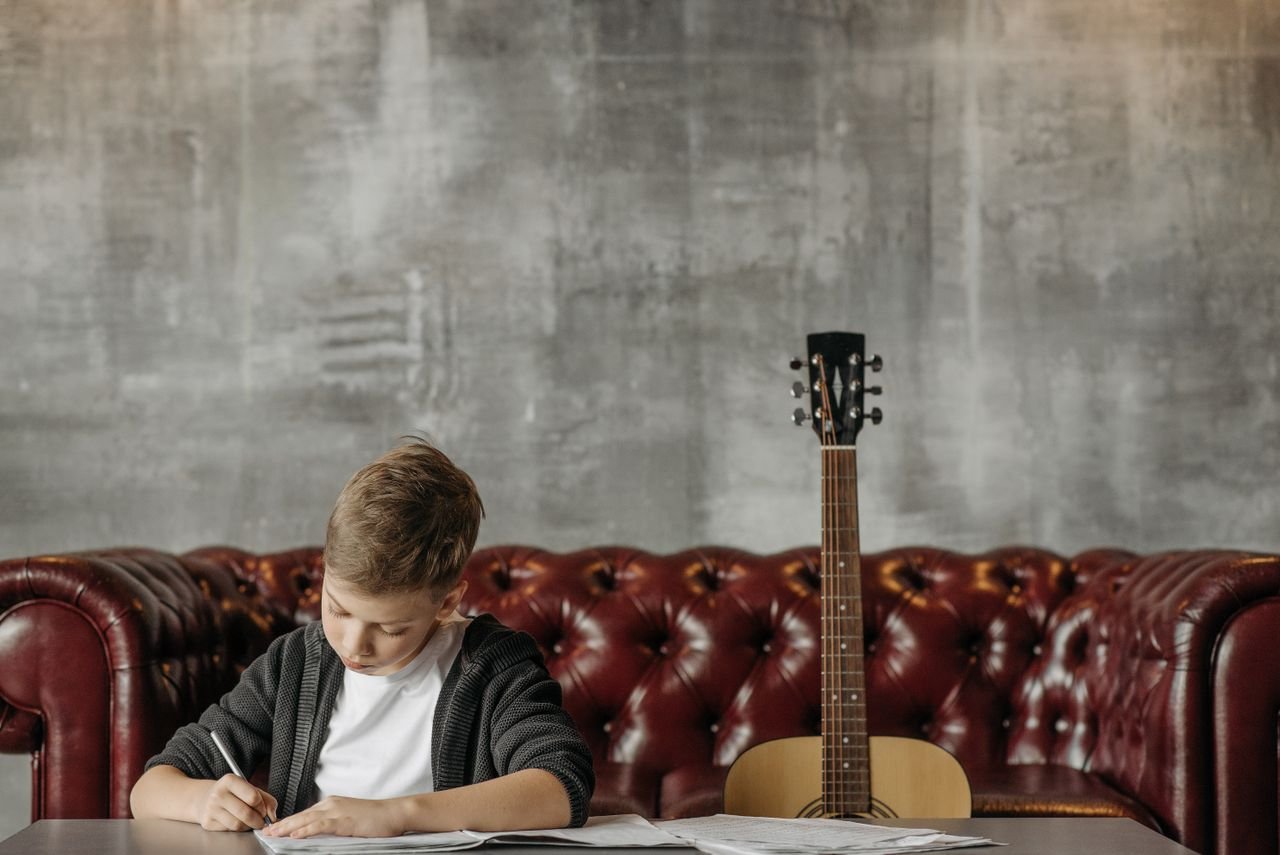 Young boy writing music notes with a guitar beside him on a red sofa.