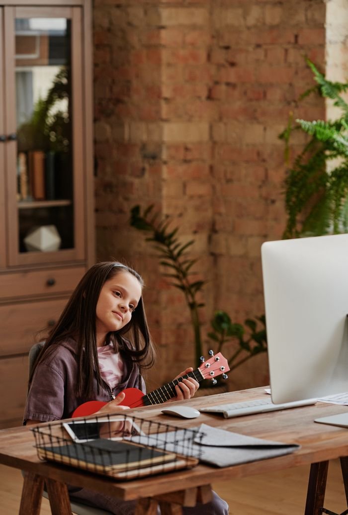 Young girl learning to play ukulele in a cozy home setting during an online lesson.