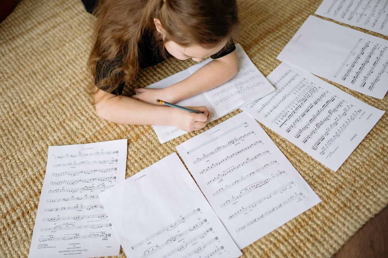 10 Ways to Motivate Teenage Music Students A young girl lying on a carpet writing music notes surrounded by sheets, enjoying a creative moment at home.